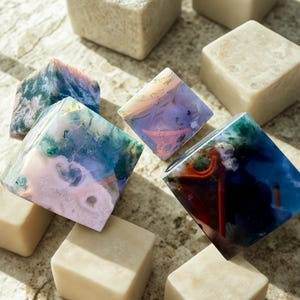 May include: An assortment of decorative stone cubes and diamond-shaped stones. The cubes are a light beige color, while the diamond-shaped stones display a variety of colors, including blue, green, pink, and orange. The stones are arranged on a textured surface.