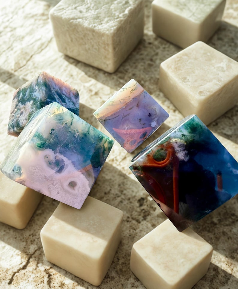 May include: An assortment of decorative stone cubes and diamond-shaped stones. The cubes are a light beige color, while the diamond-shaped stones display a variety of colors, including blue, green, pink, and orange. The stones are arranged on a textured surface.