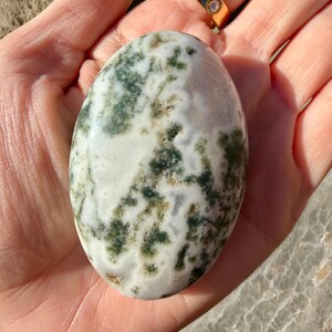 May include: An oval-shaped stone with a white base and green and brown patterns. The stone is held in a hand, and a gold ring with three small diamonds is visible in the background. The stone's surface has a natural, organic appearance.