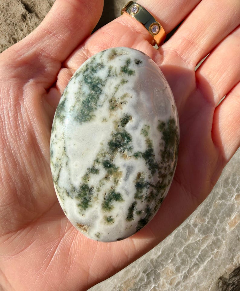 May include: An oval-shaped stone with a white base and green and brown patterns. The stone is held in a hand, and a gold ring with three small diamonds is visible in the background. The stone's surface has a natural, organic appearance.
