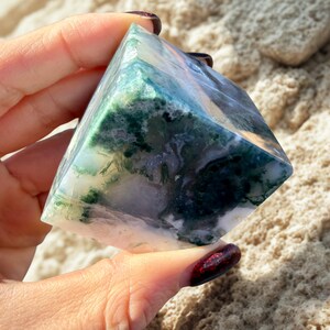May include: A polished, pyramid-shaped gemstone with a mix of green, white, and blue hues. The stone has a translucent quality, revealing intricate patterns within. The pyramid is held in a hand, showcasing its size and shape.