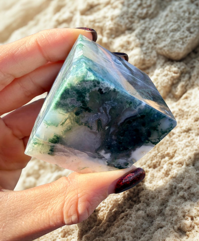 May include: A polished, pyramid-shaped gemstone with a mix of green, white, and blue hues. The stone has a translucent quality, revealing intricate patterns within. The pyramid is held in a hand, showcasing its size and shape.