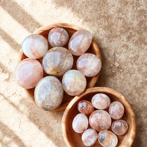 May include: Two wooden bowls filled with polished, translucent gemstones. The stones are primarily clear with internal inclusions of pink, orange, and brown. The bowls sit on a textured, light brown surface, with shadows from overhead.