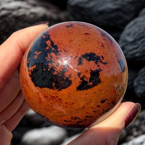 May include: A polished, spherical gemstone with a vibrant orange and black mottled pattern. The orb is held in a hand, showcasing its smooth, reflective surface. The stone's colors and unique markings create a striking visual effect.