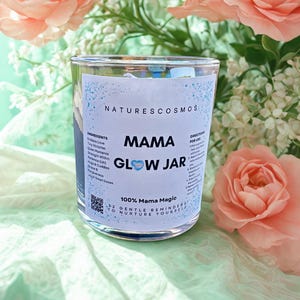 May include: A clear glass jar labeled "MAMA GLOW JAR" with the text "NATURESCOSMOS." The jar contains small items and has a blue and white label with the words "100% Mama Magic" and "32 Gentle Reminders to Nurture Yourself." The background features pink flowers.