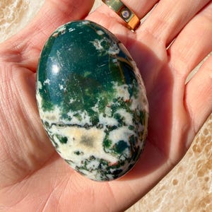 May include: A polished, oval-shaped gemstone with a mix of deep green, white, and cream colors. The stone has a marbled appearance with intricate patterns. A gold ring with three small clear stones is visible in the background.