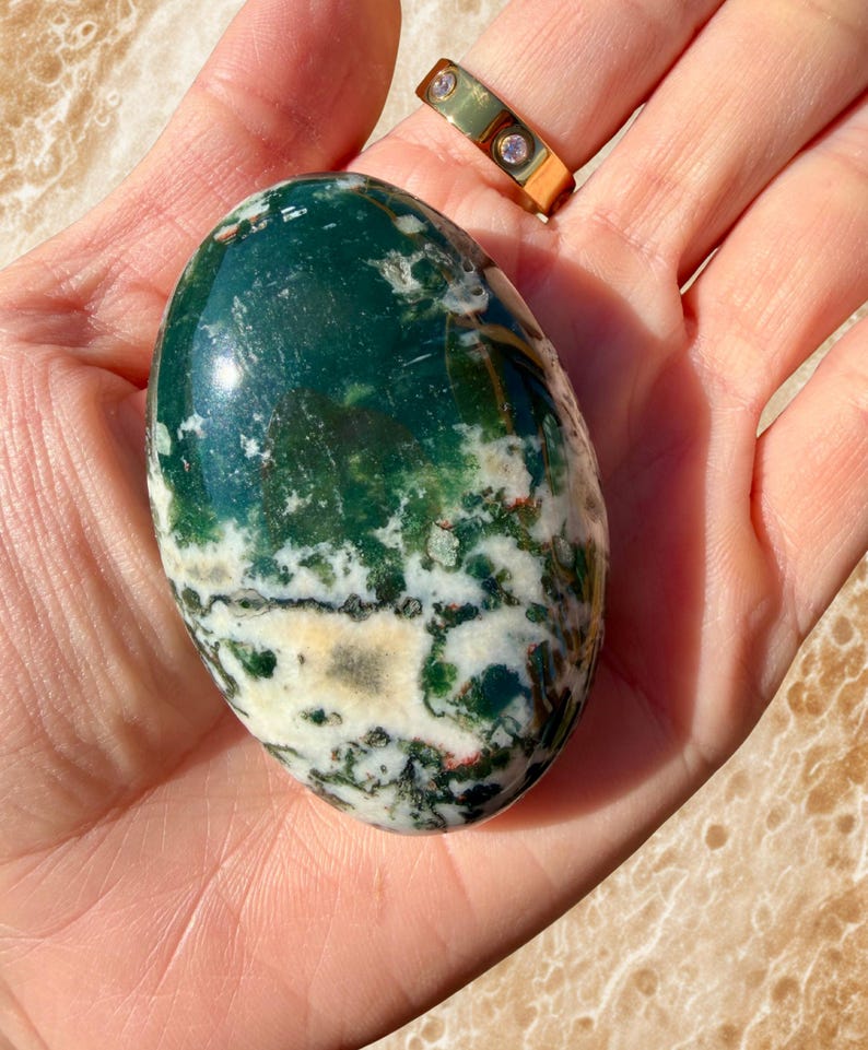 May include: A polished, oval-shaped gemstone with a mix of deep green, white, and cream colors. The stone has a marbled appearance with intricate patterns. A gold ring with three small clear stones is visible in the background.