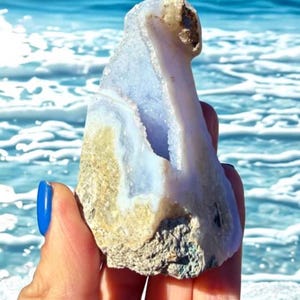 May include: A natural, raw blue lace agate crystal geode. The stone has a light blue interior with white and tan accents. The geode is held against a backdrop of the ocean, with blue water and white waves.