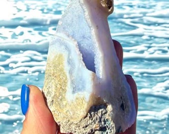 May include: A natural, raw blue lace agate crystal geode. The stone has a light blue interior with white and tan accents. The geode is held against a backdrop of the ocean, with blue water and white waves.