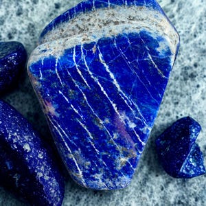 May include: A collection of polished lapis lazuli stones. The largest stone is a deep blue with white veining and a band of gray and white at the top. Smaller stones are scattered around the main piece, all displaying the same rich color and patterns.