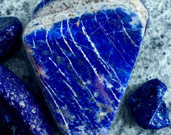 May include: A collection of polished lapis lazuli stones. The largest stone is a deep blue with white veining and a band of gray and white at the top. Smaller stones are scattered around the main piece, all displaying the same rich color and patterns.
