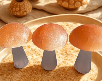May include: Three orange mushroom-shaped objects with gray stems are arranged on a decorative plate. The plate has a textured surface and a light brown color. Other decorative objects, including a small vase with yellow flowers, are in the background.