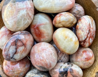 May include: A collection of polished, oval-shaped stones in shades of cream, peach, and reddish-brown. The stones have a smooth, glossy surface with natural veining and patterns. The stones are arranged in a wooden bowl, creating a natural and earthy aesthetic.