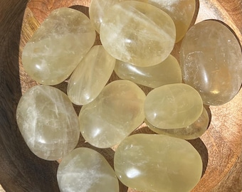 May include: A collection of smooth, oval-shaped yellow calcite stones. The stones vary in size and have a translucent quality, with subtle internal patterns. They are arranged in a wooden bowl, creating a natural and earthy aesthetic.