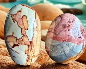 May include: Two polished, oval-shaped stones with unique patterns. One features a cream base with brown and red veining, the other a mix of pink, blue, and gray. They rest on textured, orange-brown rocks, with a blurred ocean backdrop.