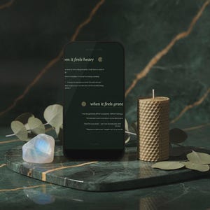 以下が含まれることがあります： when it feels heavyおよびwhen it feels grateというタイトルのテキストが表示されたスマートフォン。ハニカム構造の円筒形蜜蝋キャンドル、虹色に輝くムーンストーン、緑色のユーカリの葉が、濃い緑色の大理石の表面に配置されています。