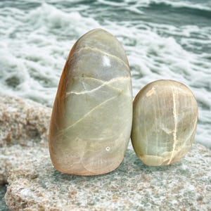 May include: Two smooth, oval-shaped stones in shades of green and tan rest on a textured rock surface. The larger stone is upright, while the smaller one is rounded. The background features a blurred view of ocean waves.