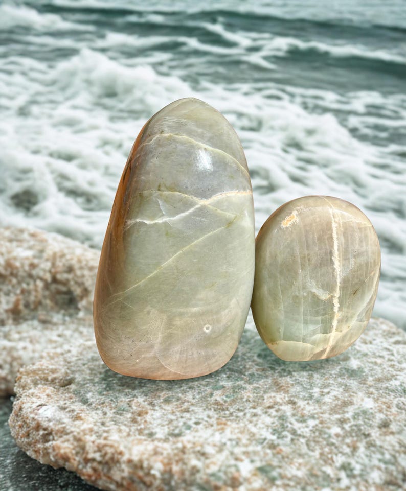 May include: Two smooth, oval-shaped stones in shades of green and tan rest on a textured rock surface. The larger stone is upright, while the smaller one is rounded. The background features a blurred view of ocean waves.