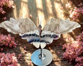 May include: A decorative butterfly sculpture with silver wings and a brown body, mounted on a silver stand. The wings have a detailed, textured design. Pink flowers and wooden planks form the background.
