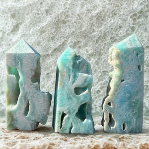 May include: Three turquoise-colored crystal obelisks with carved, textured surfaces. The obelisks have a mix of light blue, white, and green hues. The crystals are displayed on a textured, light brown surface, with a white, textured background.