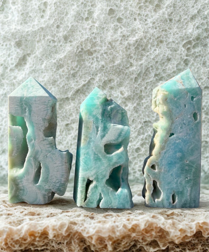 May include: Three turquoise-colored crystal obelisks with carved, textured surfaces. The obelisks have a mix of light blue, white, and green hues. The crystals are displayed on a textured, light brown surface, with a white, textured background.