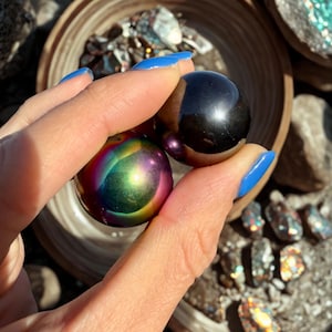 May include: A hand holds two polished spheres: one iridescent with a rainbow sheen, the other a dark, reflective black. The background includes a wooden bowl with crystals and rocks, suggesting a focus on gemstones or decorative objects.