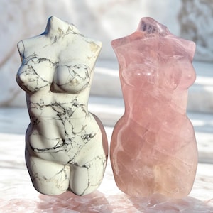 May include: Two carved stone torso sculptures. One is white with gray veining, the other is a translucent pink. The sculptures are of a female form, with defined breasts and a smooth, polished finish. They are displayed on a pink surface.