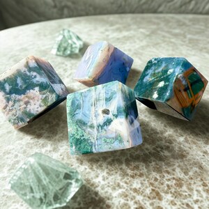 May include: An assortment of polished gemstone cubes in shades of blue, green, and brown. Two clear, faceted crystal shapes are also present. The gemstones have unique patterns and textures, showcasing natural variations.