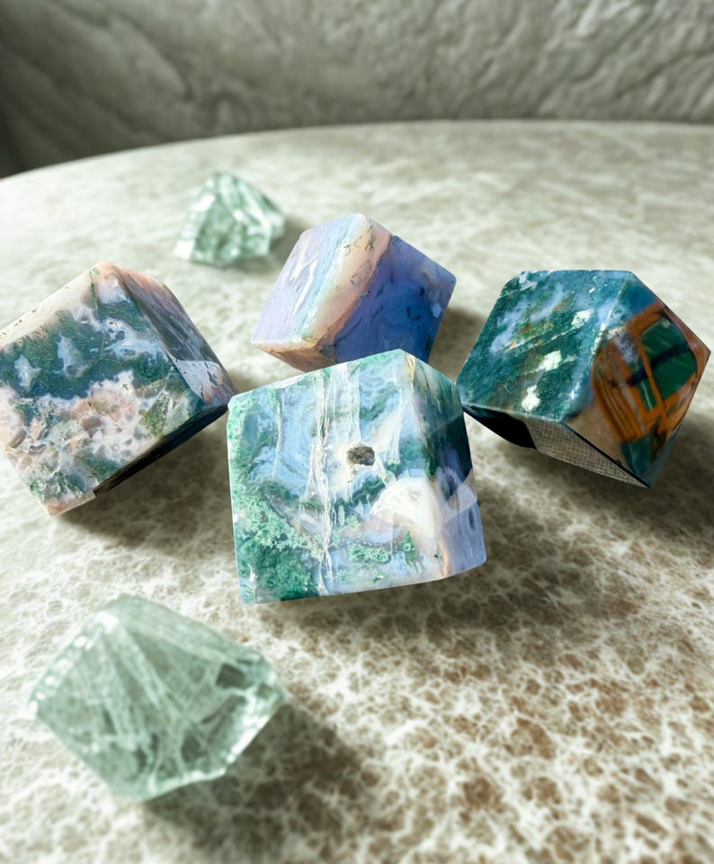 May include: An assortment of polished gemstone cubes in shades of blue, green, and brown. Two clear, faceted crystal shapes are also present. The gemstones have unique patterns and textures, showcasing natural variations.