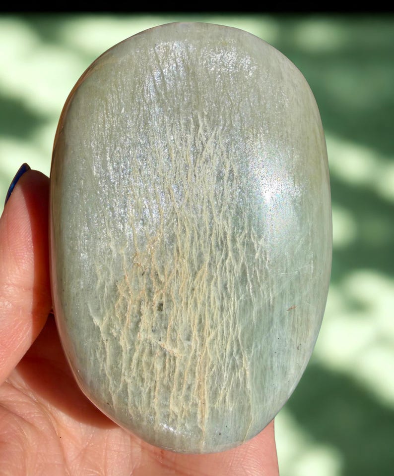 May include: A smooth, oval-shaped stone with a pale green hue and a textured surface. The stone features light beige vein-like patterns across its surface. The stone is held in a hand, with a blurred green background.