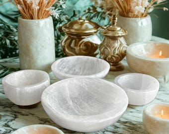May include: A collection of white selenite bowls and candle holders. The bowls vary in size and shape, with a natural, translucent appearance. Gold-colored decorative jars and vases with dried flowers are in the background.