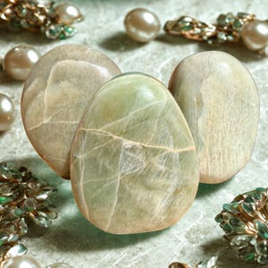 May include: Three polished, oval-shaped stones in shades of green and beige, with visible veining. The stones are arranged on a textured surface, surrounded by pearls and gold-toned jewelry with green accents.