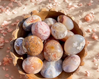 May include: A wooden bowl filled with polished gemstones in shades of pink, brown, and white. The bowl sits on a surface scattered with pink flower petals and a rose.