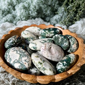 May include: A wooden bowl filled with polished moss agate stones. The stones are oval-shaped, with a mix of white, green, and brown patterns. The bowl has a scalloped edge and is made of light-colored wood.
