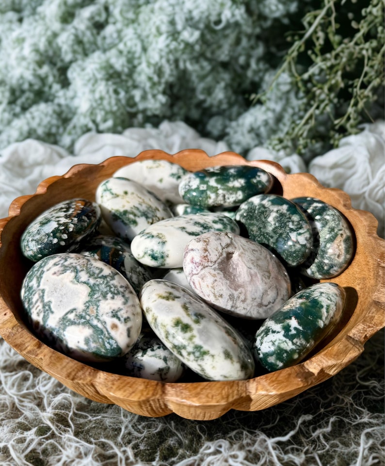 May include: A wooden bowl filled with polished moss agate stones. The stones are oval-shaped, with a mix of white, green, and brown patterns. The bowl has a scalloped edge and is made of light-colored wood.