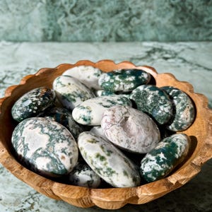 May include: A wooden bowl filled with polished stones. The stones are oval-shaped and feature a mix of white, green, and gray patterns. The bowl has a scalloped edge and sits on a marble surface.