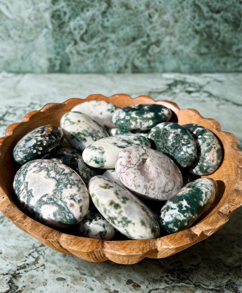 May include: A wooden bowl filled with polished stones. The stones are oval-shaped and feature a mix of white, green, and gray patterns. The bowl has a scalloped edge and sits on a marble surface.