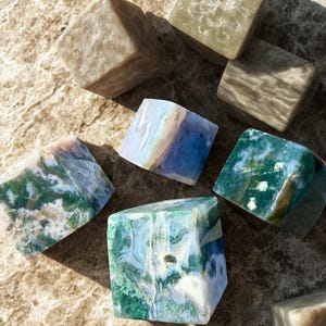 May include: An assortment of square-cut gemstones in various colors, including green, blue, and beige. The gemstones are arranged on a light brown, marble-like surface, with natural light casting shadows.
