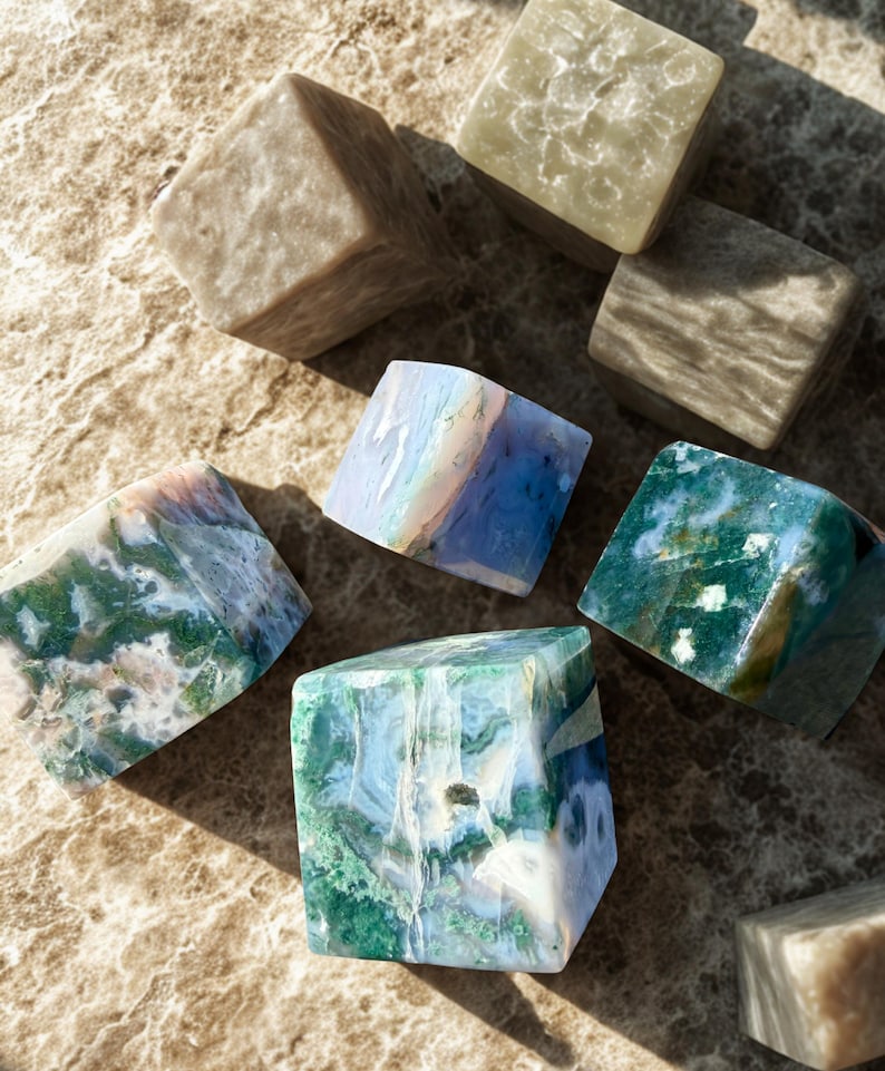 May include: An assortment of square-cut gemstones in various colors, including green, blue, and beige. The gemstones are arranged on a light brown, marble-like surface, with natural light casting shadows.