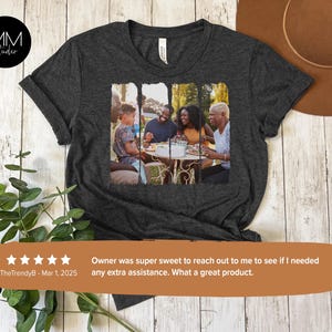 May include: A dark gray t-shirt with a photo of a family of four sitting at a table outdoors. The photo is printed in a rectangular shape on the front of the shirt.