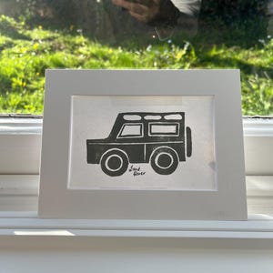 Pode incluir: Impressão emoldurada em preto e branco de um Land Rover. A obra de arte apresenta uma ilustração estilizada do veículo com rodas redondas e um pneu sobressalente. A impressão está em uma moldura branca, exibida em uma janela branca.
