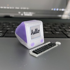 May include: A miniature computer with a purple top and white body, a small keyboard, and a mouse. The computer screen displays the word "hello" in cursive. The items are on a laptop's surface.