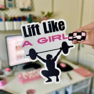 May include: A white sticker with a black silhouette of a person lifting a barbell. The text "Lift Like A Girl" is written in black and pink letters.