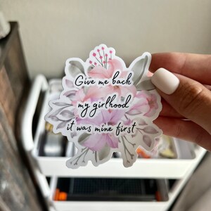Può includere: Un adesivo bianco con un design floreale e il testo "Give me back my girlhood it was mine first".