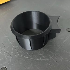 以下が含まれることがあります： 黒色の3Dプリントされた車のカップホルダー。カップホルダーは円形の開口部と、湾曲したエッジを持つ長方形のベースを備えています。デザインには側面に垂直スロットが含まれています。このアイテムはプラスチック製です。