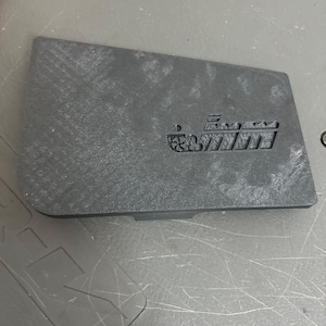 Puede incluir: Una tapa de plástico negro con una imagen en relieve de un barco. El barco tiene un contorno blanco y un pequeño círculo en la parte delantera.