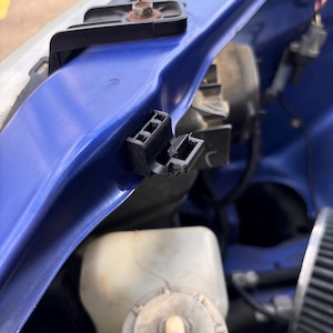 Op de afbeelding: Close-up van een zwarte plastic connector met meerdere pinnen, bevestigd aan een blauw auto-onderdeel. De connector is zichtbaar tegen een blauwe achtergrond.