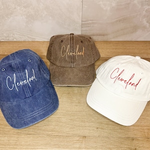 Puede incluir: Tres gorras de béisbol con la palabra "Cleveland" bordada. Las gorras son marrón, blanca y azul.
