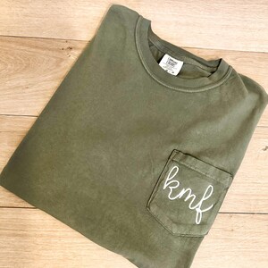 May include: A green t-shirt with a pocket on the front. The pocket has the letters "kmf" embroidered on it in white thread.