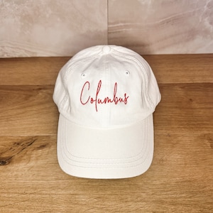 Może przedstawiać: Biała czapka bejsbolówka z haftowanym czerwonym napisem "Columbus".